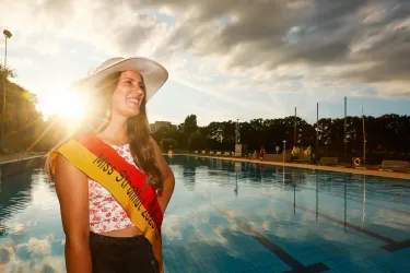 Eine lachende Frau steht am Rand eines Schwimmbades und trägt einen Sonnenhut. Sie hat ein Banner mit der Aufschrift „Miss Schönheiten 2023“.