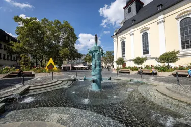Ein schöner Platz mit einem Brunnen im Mittelpunkt. Im Hintergrund sind Bäume und ein historisches Gebäude zu sehen.