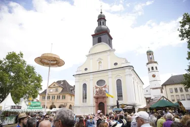 Ein belebter Marktplatz mit einer großen Kirche im Hintergrund. Menschen stehen zusammen und genießen die Atmosphäre.