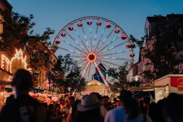Ein belebter Jahrmarkt bei Dämmerung mit einem großen Riesenrad im Hintergrund. Viele Menschen genießen die festliche Atmosphäre und die bunten Lichter.