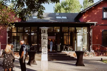 Ein modernes Gebäude mit großen Fenstern und einer einladenden Eingangstür. Im Vordergrund stehen einige Personen und Stehtische.