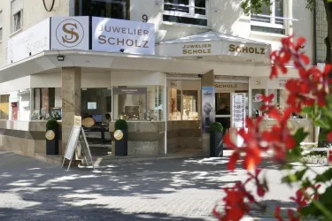 Ein Juweliergeschäft mit der Aufschrift "Juwelier Scholz" an einer Straße. Im Vordergrund blühen rote Blumen, die das Bild verschönern.