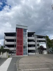 Ein modernes, mehrstöckiges Gebäude mit einer roten Fassade und Glasfront. Der Himmel ist bewölkt und es ist eine Parkfläche im Vordergrund zu sehen.