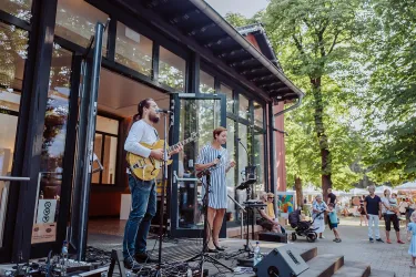 Eine Live-Band spielt draußen vor einem Gebäude. Menschen stehen um sie herum und genießen die Musik.