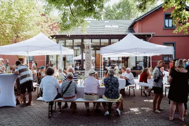 Eine fröhliche Veranstaltung im Freien mit mehreren Tischen und Sonnenschirmen. Viele Menschen sitzen zusammen und genießen das Beisammensein.