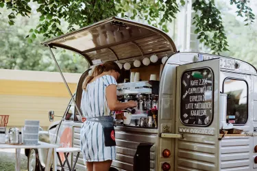 Ein gemütlicher Kaffee-Wagen mit einer Barista, die Kaffee zubereitet. Im Hintergrund sind Bäume und eine entspannte Atmosphäre zu sehen.