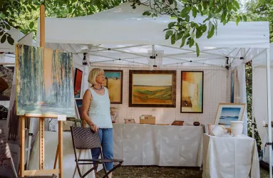 Eine Künstlerin steht in ihrem Stand auf einem Kunstmarkt. Im Hintergrund sind verschiedene Gemälde und Kunstwerke ausgestellt.