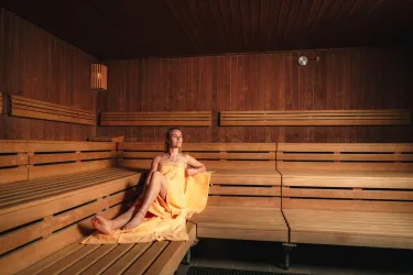Eine Frau in einem eleganten orangefarbenen Kleid sitzt entspannt in einer Holzsauna. Die Wände und Bänke sind aus hellem Holz, und es gibt sanftes Licht im Raum.