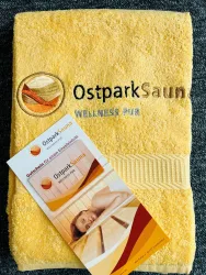 Ein gelbes Handtuch mit dem Aufdruck "OstparkSaun" und dem Text "WELLNESS PUR". Daneben liegt ein Informationsflyer über die OstparkSauna.