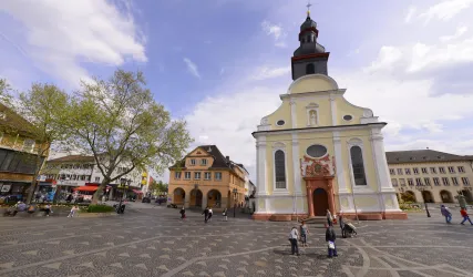 Ein schöner Platz mit einer Kirche im Vordergrund und ruhigen Gebäuden im Hintergrund. Menschen schlendern auf dem Platz und genießen die Atmosphäre.