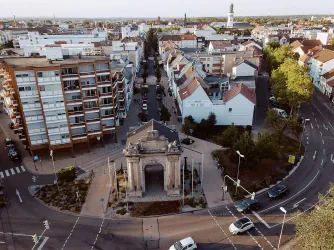 Eine Luftaufnahme einer Stadt mit Wohnhäusern und einer zentralen Rundung. In der Mitte steht ein historisches Bauwerk, das als Tor dient.