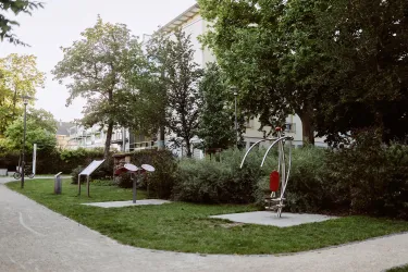Ein ruhiger Park mit vielen Bäumen und einer Grünfläche. Im Vordergrund sind verschiedene Spiel- und Trainingsgeräte zu sehen.