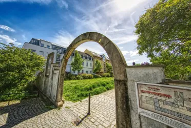 Ein schöner Bogen aus Stein führt in einen grünen Garten. Im Hintergrund sind Häuser und blauer Himmel zu sehen.
