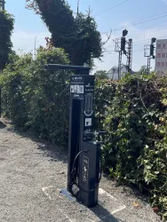 Eine öffentliche E-Ladestation für Elektroautos steht auf einem gepflasterten Bereich. Im Hintergrund sind Gleise und ein bewachsener Zaun zu sehen.