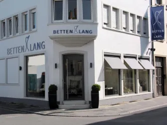 Ein modernes Geschäft mit dem Namen "BETTEN LANG" an einer Straßenecke. Die Fassade ist hell und hat große Fenster mit Fensterläden.