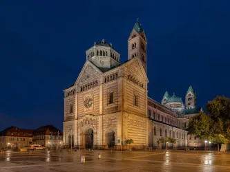 Im Vordergrund des Bildes ist der gepflasterte Platz des Speyerer Doms. Im Zentrum steht der Speyerer Dom bei Nacht, dieser wird beleuchtet.