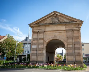 Ein historisches Tor mit einer dekorativen Fassade. Rund um das Tor blühen bunte Blumen und es ist ein klarer blauer Himmel zu sehen.