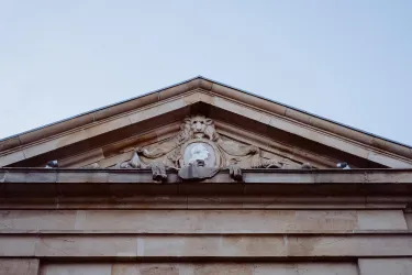 Ein historisches Gebäude mit kunstvollen Verzierungen an der Dachspitze. Ein Löwenkopf ist zentral zwischen den dekorativen Elementen zu sehen.