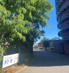 Ein schmaler Weg mit Bäumen und einem Parkplatz-Schild. Im Hintergrund sieht man ein modernes Gebäude.
