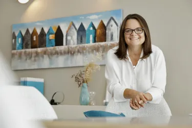 Eine lächelnde Frau mit Brille steht in einem modernen Raum. Im Hintergrund hängt ein Bild mit bunten kleinen Häusern.