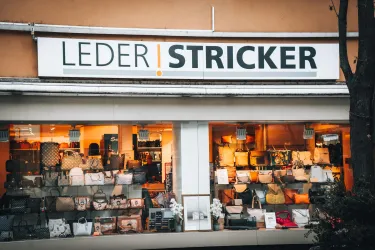 Ein Schaufenster eines Lederwarenladens mit verschiedenen Taschen und Accessoires. Das Geschäft hat eine auffällige Beschilderung mit dem Namen "LEDER STRICKER".