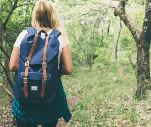 Eine Person mit einem blauen Rucksack wandert einen Waldweg entlang. Die Umgebung ist grün und von Bäumen umgeben.