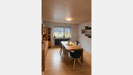 Ein helles Esszimmer mit einem holzfarbenen Tisch und modernen Stühlen. Große Fenster sorgen für viel Tageslicht und eine einladende Atmosphäre.