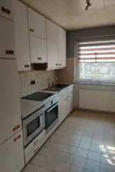 Eine moderne Küche mit weißen Schränken und einem Fenster, das Tageslicht hereinlässt. Es gibt einen Herd, einen Backofen und ein Spülbecken.