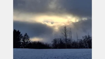 Ein winterlicher Himmel mit dichten, grauen Wolken und einem schimmernden Lichtstrahl. Im Vordergrund liegt eine schneebedeckte Landschaft mit einzelnen Bäumen.