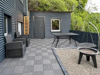 Eine moderne Terrasse mit einem Tisch und Stühlen aus Rattan. Im Hintergrund befindet sich ein Grillplatz mit Kies und einer Feuerstelle.
