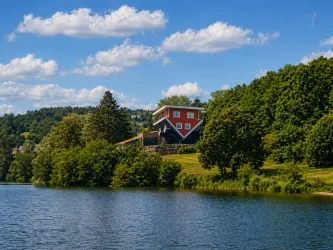 Ein malerisches Haus auf einem Hügel mit Blick auf einen ruhigen See. Umgeben von üppigem Grün und einem blauen Himmel mit weißen Wolken.