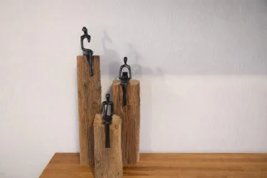 Drei moderne Figuren aus Metall stehen auf Holzblöcken. Sie sind minimalistischer Art und strahlen eine ruhige Atmosphäre aus.