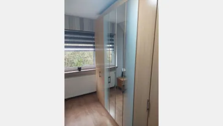 Ein heller Raum mit einem Kleiderschrank und einem Fenster. Die Wände sind hell gestrichen, und es gibt einen kleinen Tisch.