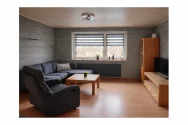 Ein gemütliches Wohnzimmer mit einem dunklen Sofa und einem Couchtisch. Große Fenster mit Jalousien sorgen für viel Licht im Raum.