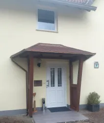 Ein Eingangsbereich mit einer weißen Tür und einem Vordach aus Holz. Die Wand ist gelb gestrichen und es stehen einige Pflanzen in Töpfen.