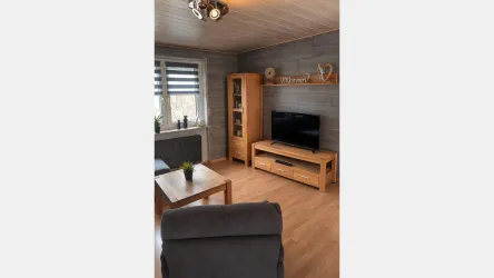 Ein gemütliches Wohnzimmer mit einer hellen Holzverkleidung. Es gibt ein Fernsehmöbel, einen Tisch und bequeme Stühle.