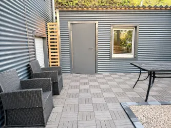 Eine moderne Terrasse mit einem Tisch und zwei Stühlen aus Rattan. Im Hintergrund befindet sich eine graue Tür und ein Fenster in einer wellenförmigen Wand.