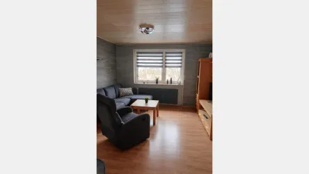 Ein gemütliches Wohnzimmer mit einer blauen Couch und einem Sessel. Es gibt einen Tisch und große Fenster mit Jalousien, die für Tageslicht sorgen.