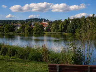 Ein ruhiger See, umgeben von üppigem Grün und Bäumen. Im Hintergrund sind kleine Häuser und sanfte Hügel zu sehen unter einem blauen Himmel mit weißen Wolken.