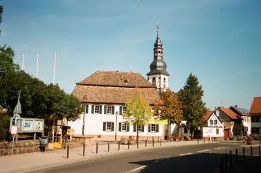 Ein malerisches Dorf mit traditionellen Gebäuden und einem hohen Kirchturm. Die Umgebung ist grün und einladend unter klarem Himmel.