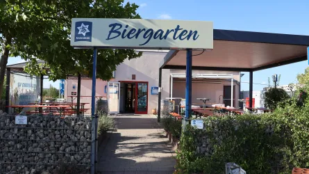 Ein Biergarten mit einem einladenden Eingang und Sitzgelegenheiten im Freien. Der Bereich ist von Pflanzen umgeben und bietet einen entspannten Ort zum Verweilen.