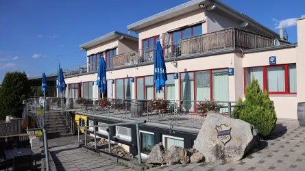 Das Bild zeigt ein modernes Gebäude mit Terrassen und blauen Schirmen. Es gibt Pflanzen und einen gepflasterten Weg vor dem Gebäude.