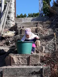 Ein Kleinkind sitzt auf einer Treppe und schaut in einen grünen Eimer. Im Hintergrund sind ein paar Pflanzen und ein blauer Himmel sichtbar.