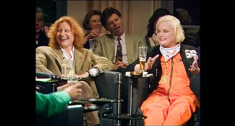 Zwei Frauen lachen und unterhalten sich fröhlich. Im Hintergrund sitzt ein Mann, der scheinbar erschöpft ist.