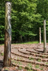 Ein labyrinthartiger Garten mit geschnitzten Holzsäulen umgeben von Bäumen. Der Boden besteht aus Steinen und Gras.