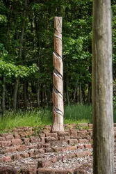 Ein kunstvoll geschnitzter Holzpfahl steht in einer grünen Waldumgebung. Der Pfahl hat eine spiralartige Verzierung und ist von einer alten Steinanlage umgeben.
