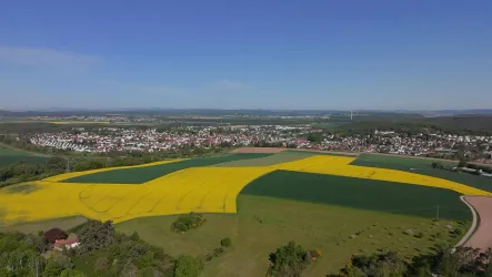 Eine weite Landschaft mit bunten Feldern, darunter leuchtend gelbe Rapsfelder und grünes Land. Im Hintergrund ist eine kleine Stadt unter einem klaren Himmel zu sehen.