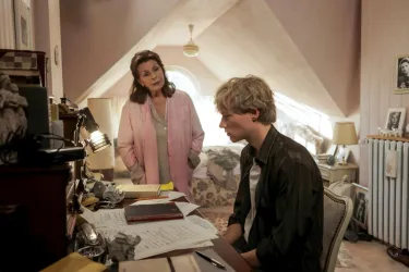 Eine Frau steht neben einem jungen Mann, der an einem Schreibtisch sitzt. Der Raum hat eine gemütliche Atmosphäre mit persönlicher Dekoration.