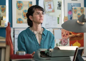Eine Frau mit Brille sitzt nachdenklich an einem Tisch. Im Hintergrund sind bunte Poster und eine Schreibmaschine zu sehen.