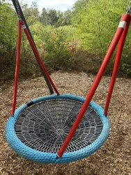 Eine große Netzschaukel in einem Spielplatzbereich, umgeben von Bäumen. Der Boden ist mit Holzspänen bedeckt.